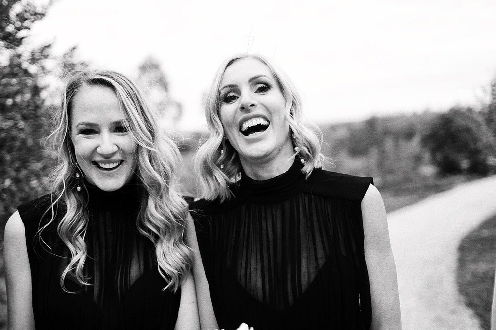Two women laughing and smiling outdoors in black sleeveless dresses, with a blurred nature background.