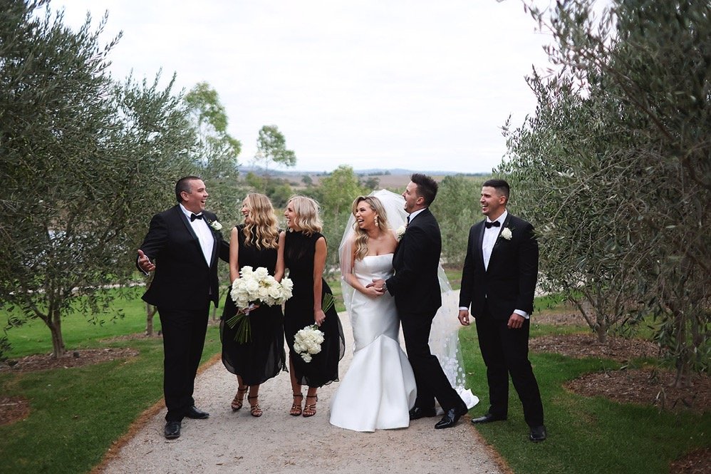 A wedding party outdoors, with a bride and groom in the center surrounded by bridesmaids and groomsmen, all smiling and chatting along a gravel path flanked by trees.