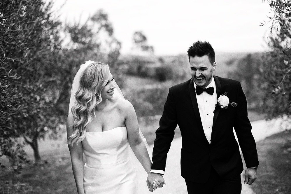 A bride and groom walking hand in hand outdoors, smiling and laughing, on their wedding day in a black-and-white photo.