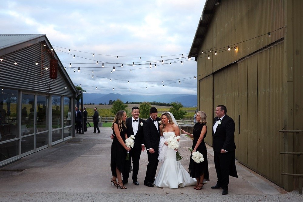 A wedding party photoshoot outdoors with a bride and groom in the center, surrounded by two men and two women, all dressed in formal attire, against a rustic setting with string lights and mountains in the background.