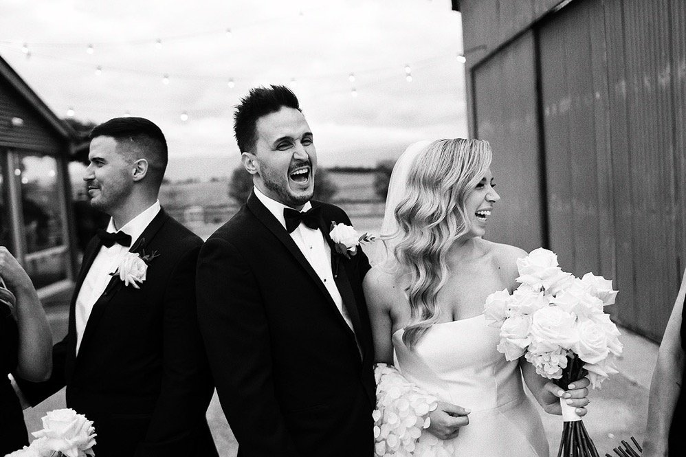 Black and white wedding photo of three people in tuxedos and a wedding dress, smiling and laughing, with a bride holding a large bouquet of white flowers.