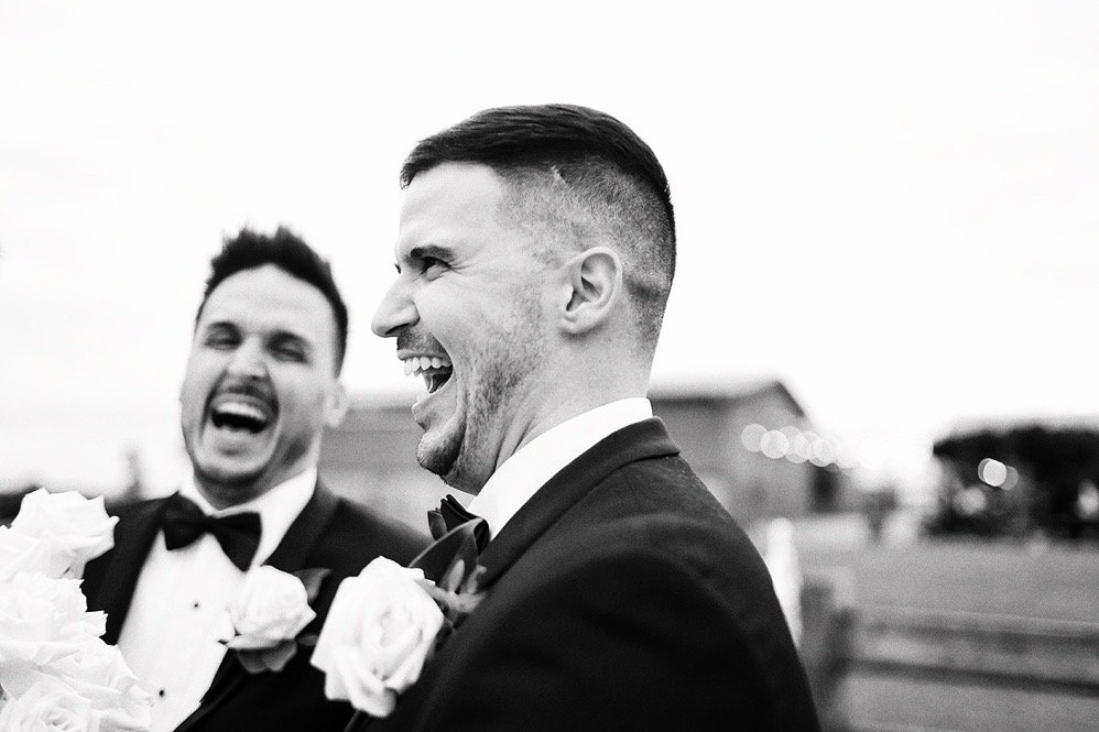Two men in tuxedos laughing and smiling at a wedding, with one holding a bouquet of roses.