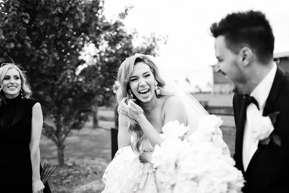 Black and white photo of a wedding scene with three people. A woman in a wedding dress is laughing, holding her face with her right hand and standing near a large bouquet of flowers. To her right, a man in a tuxedo is also laughing. On the left, anot