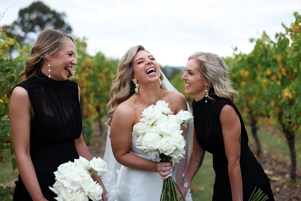 A bride in a white wedding dress holding a bouquet of white roses, laughing with two women dressed in black, also holding white rose bouquets, in a vineyard setting with green grape vines.