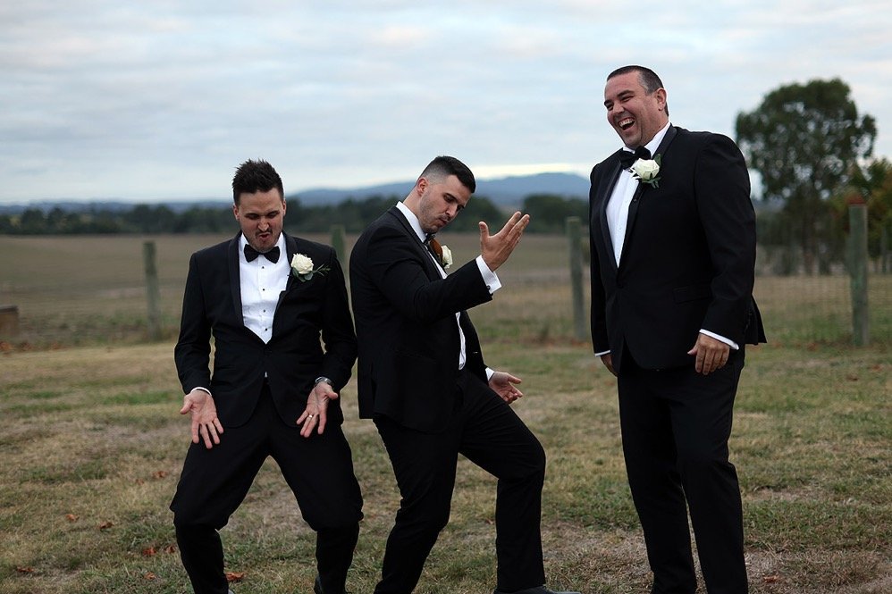 Three men in black tuxedos with white shirts and bow ties standing outdoors in a field, smiling and posing playfully for a photo, with a partly cloudy sky and trees in the background.