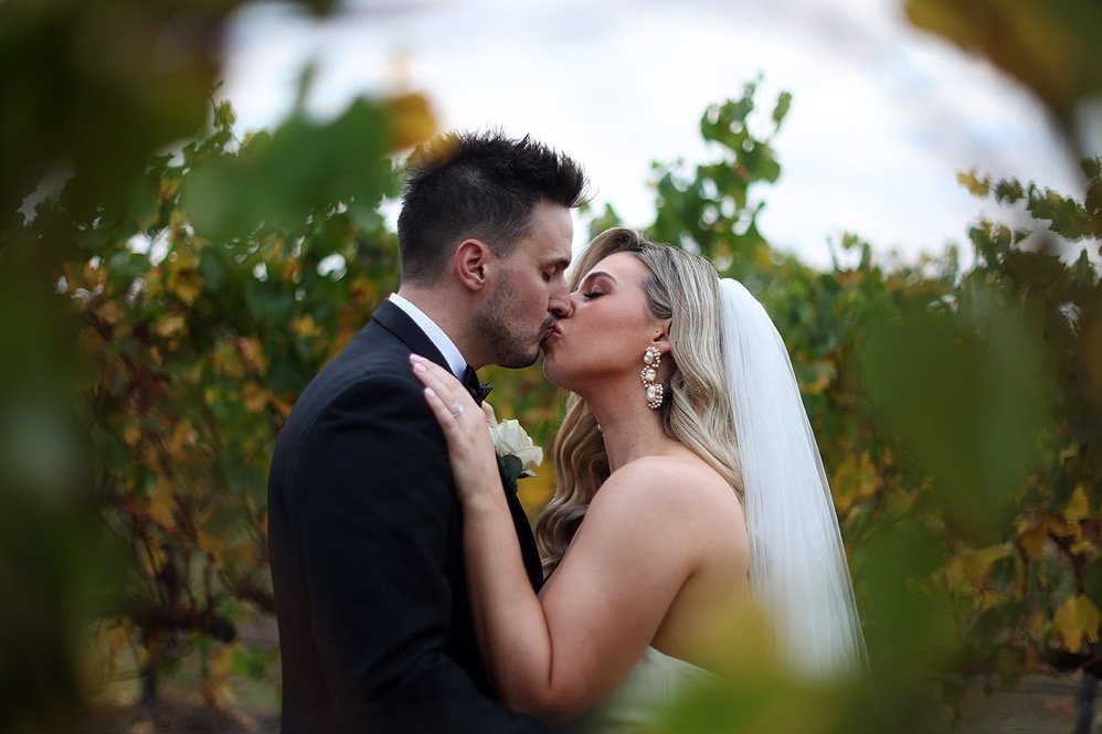 A bride and groom sharing a kiss outdoors among green foliage, dressed in wedding attire.