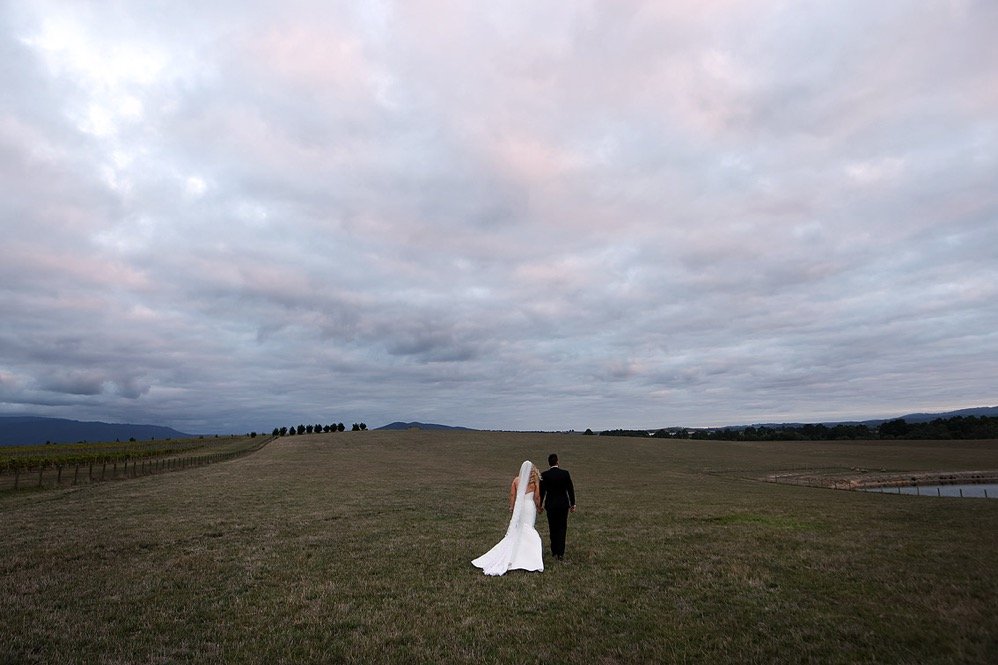 A bride and groom walking together across an open field during overcast weather.