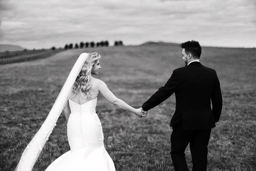 A bride and groom holding hands in an open field during their wedding photoshoot, with trees and hills in the background, black and white photography.