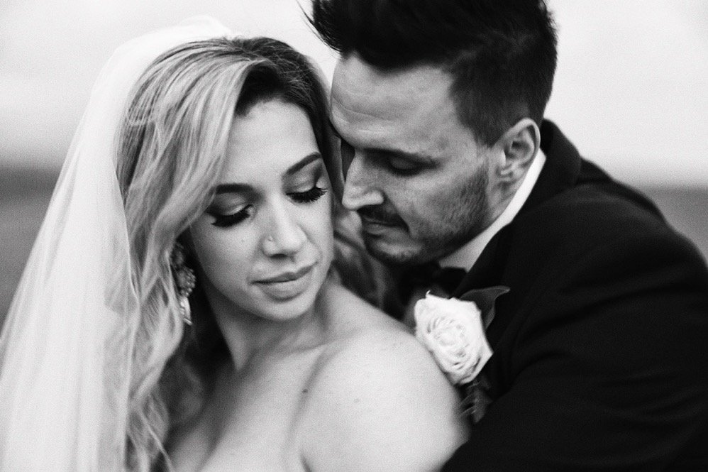 A bride and groom on their wedding day, close-up, in black and white, with the groom leaning into the bride, who is wearing a veil and jewelry.