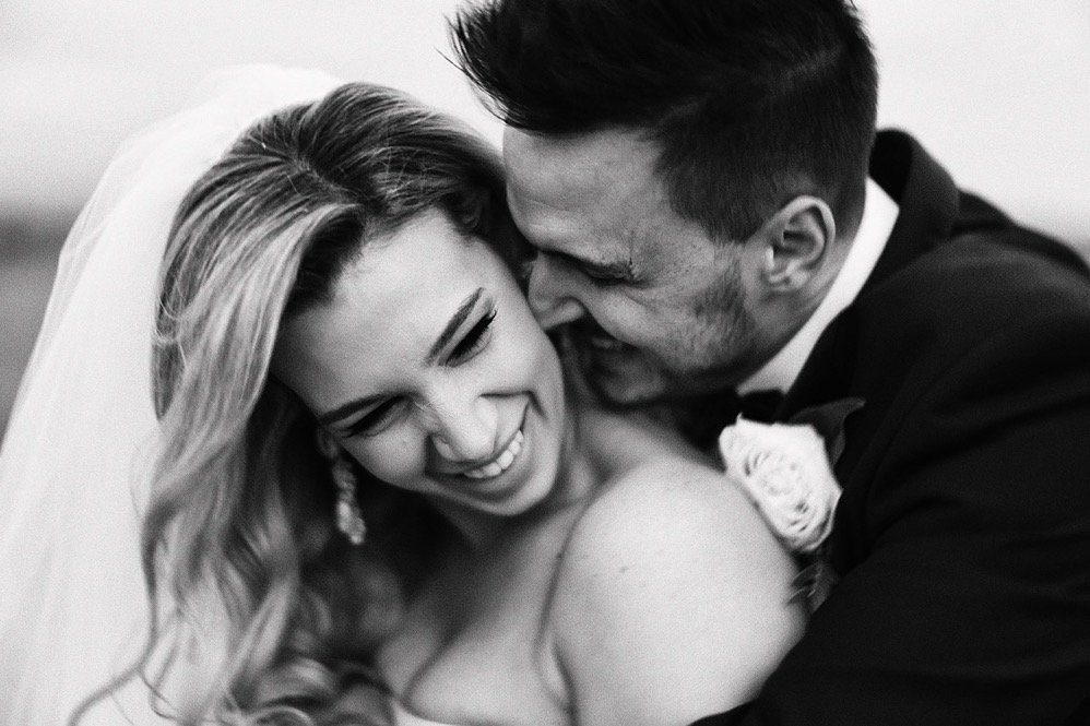 Black and white photo of a bride and groom laughing and embracing closely, wedding attire, bride with earrings and veil, groom with boutonniere.