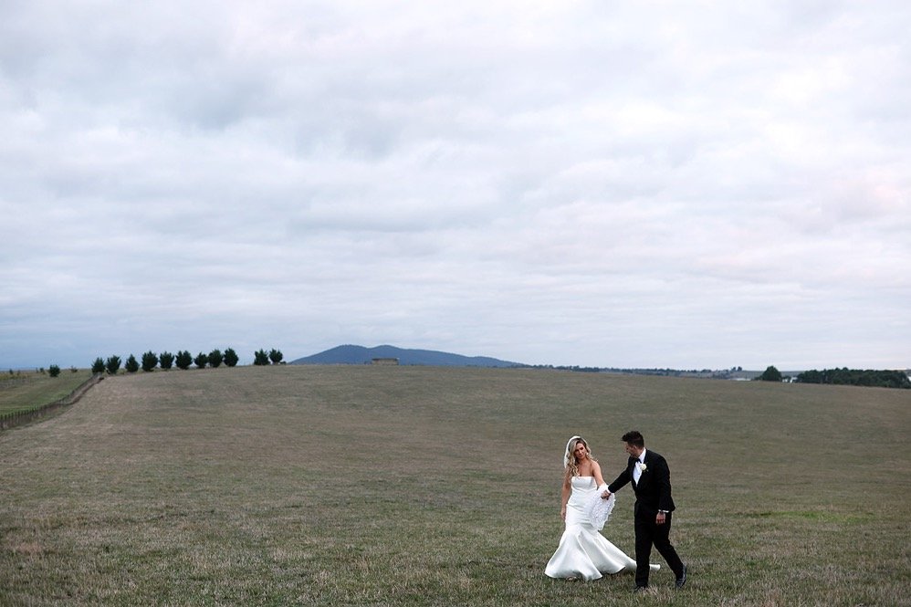 A bride and groom holding hands and walking in a vast open field with a cloudy sky in the background.