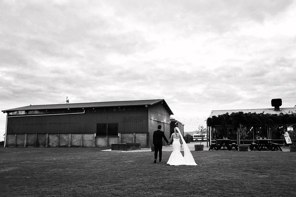 A bride and groom holding hands and walking on a grassy field near a barn and outdoor seating area at dusk.
