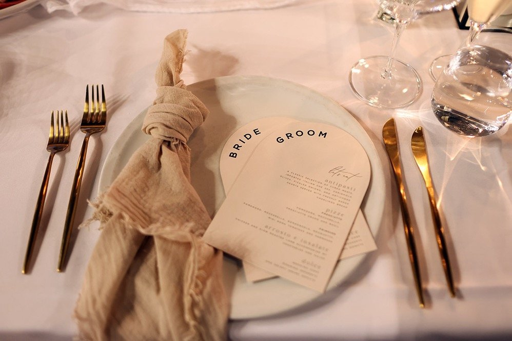 Wedding table setting with gold utensils, a cream-colored napkin tied around cutlery, a white plate with a wedding invitation card labeled 'Bride' and 'Groom,' two wine glasses, a water glass, and a glass of champagne.