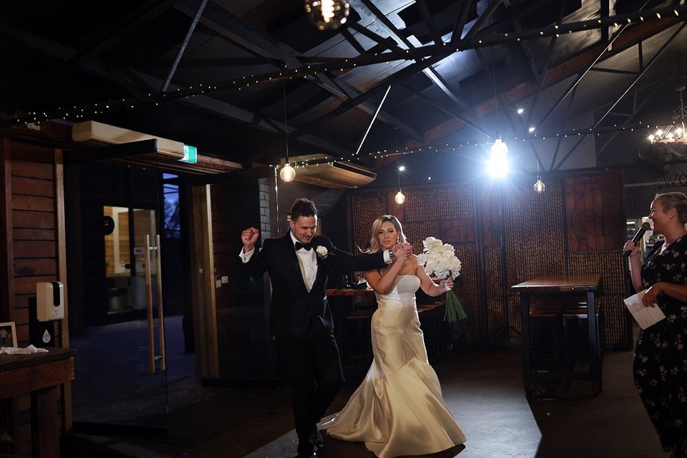 A bride and groom dancing at their wedding reception, with the bride holding a bouquet of white flowers and a woman speaking into a microphone nearby.