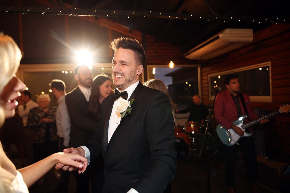 A groom in a tuxedo with a boutonniere dancing and smiling with a woman at a wedding reception, with other guests and a band in the background.