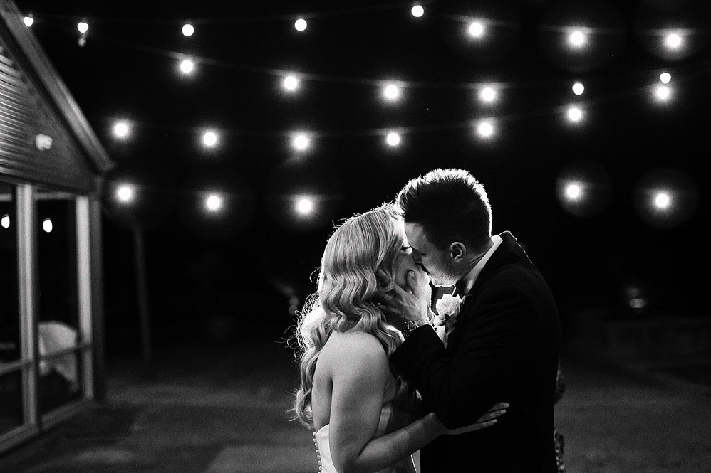 A couple sharing a kiss at night with string lights overhead, in black and white.