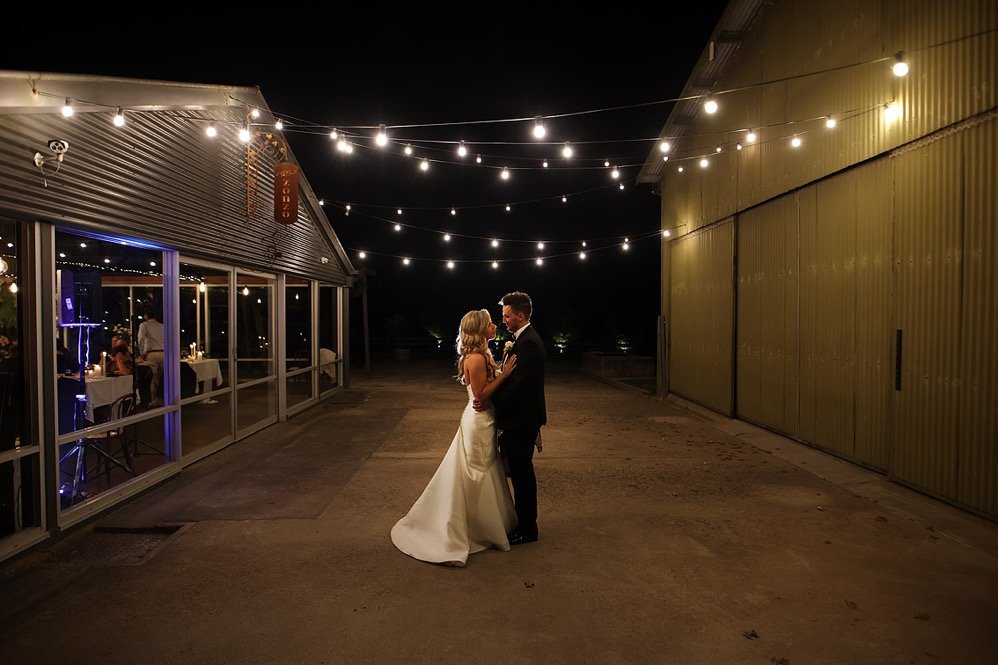 A bride and groom dancing under string lights outside at night with a yellow wall and glass-windowed building on either side.