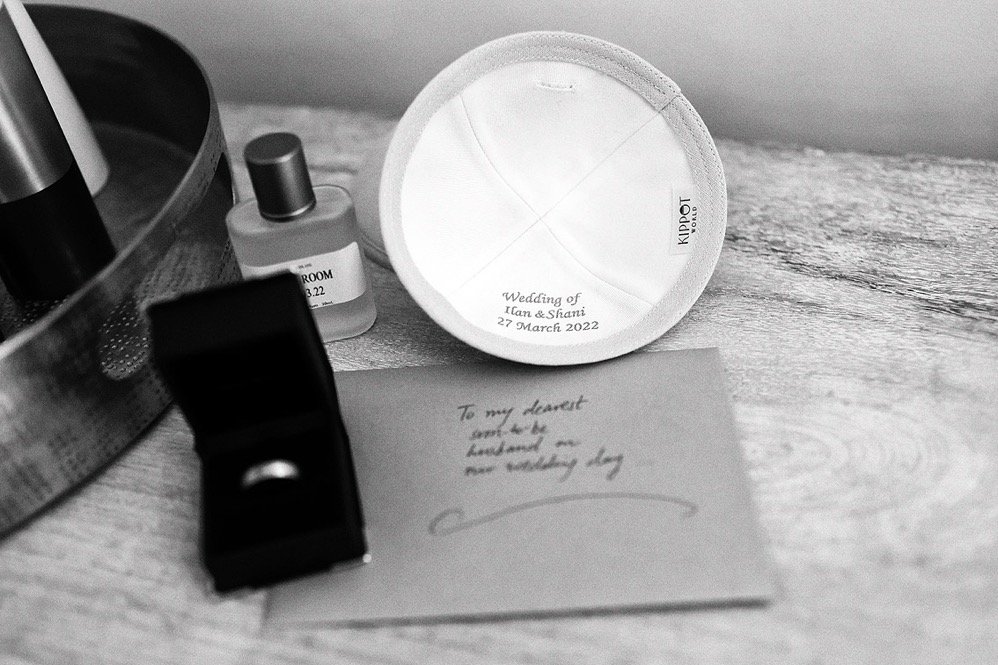 A wedding favor with the inscription "Wedding of Iman & Shani 27 March 2022", a handwritten note, a small perfume bottle, a ring in a black jewelry box, and a metallic container with other items on a wooden surface.