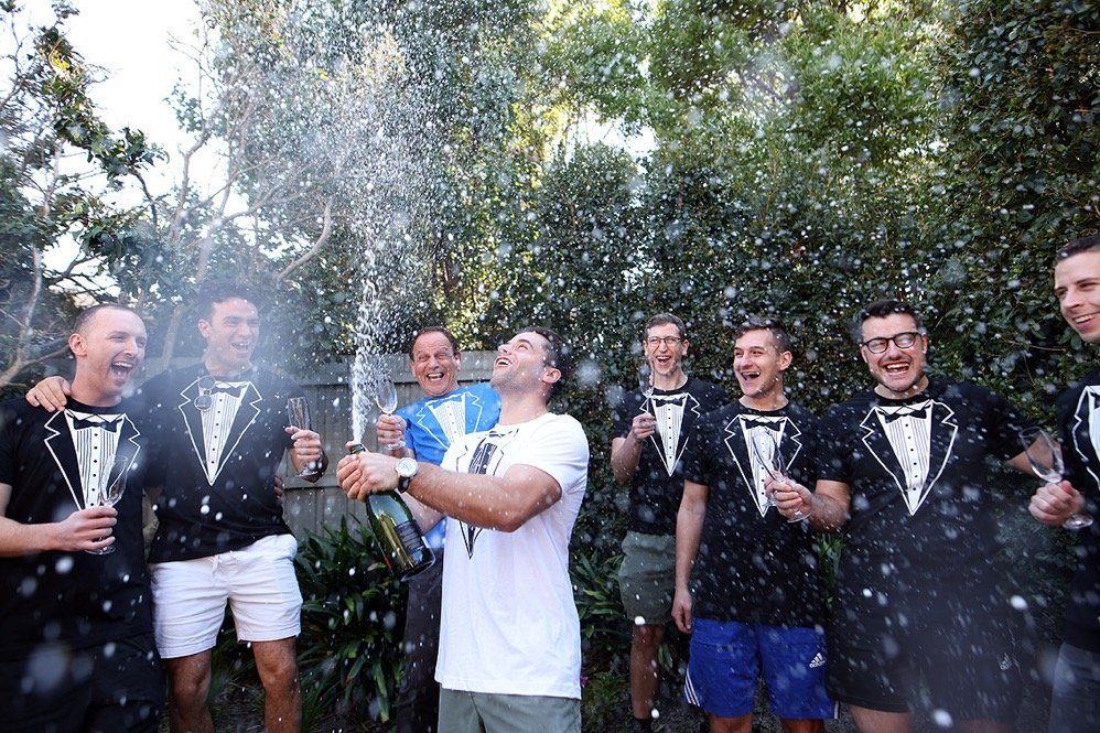 Group of friends celebrating outdoors with champagne, wearing tuxedo-themed t-shirts, as champagne sprays into the air.