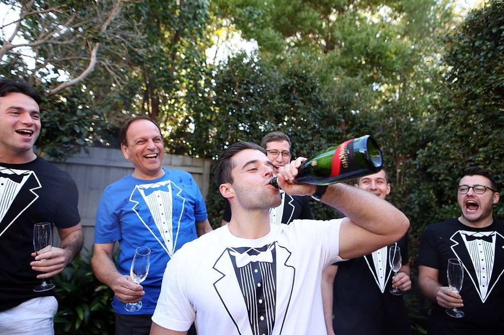 Group of men wearing tuxedo-themed T-shirts at outdoor celebration, one man drinks champagne from a bottle, others hold champagne glasses, laughing and enjoying.