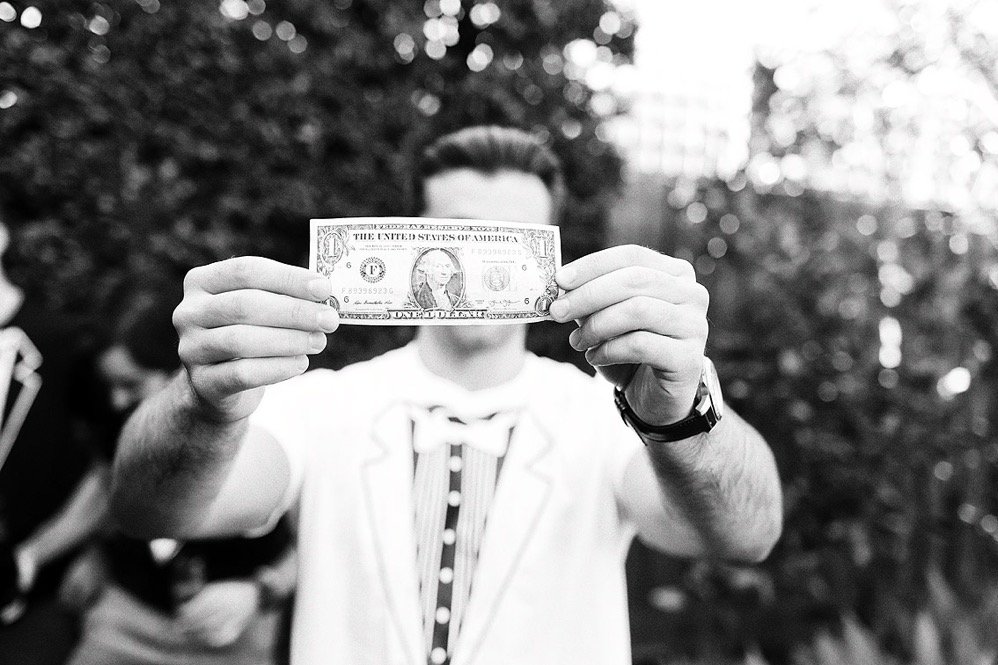 A person holding a one-dollar bill in front of their face outdoors, with other people blurred in the background.