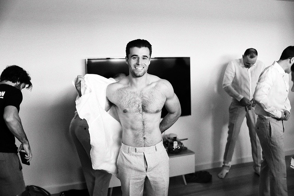 A smiling man with a shirt over his shoulder, shirtless, in a room with four other men adjusting their clothes.