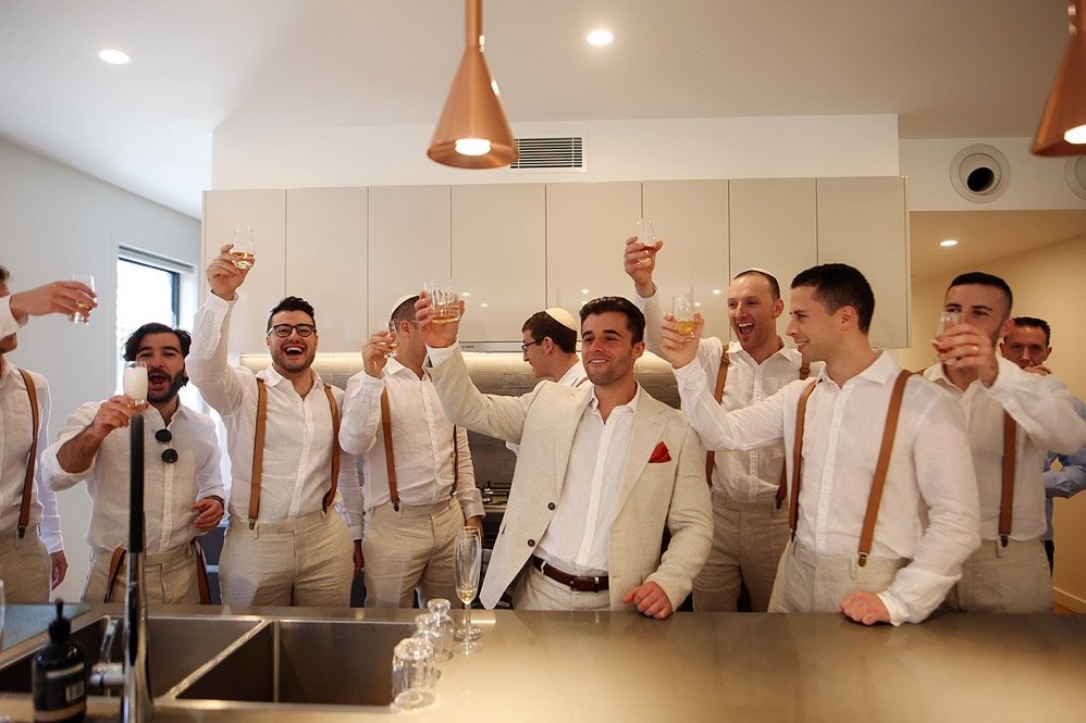 Group of men in white shirts and suspenders celebrating with drinks in a modern kitchen.