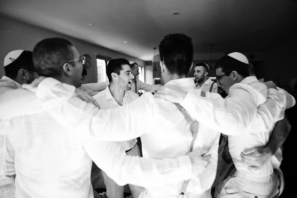 Group of men wearing white shirts and yarmulkes forming a huddle and smiling indoors.