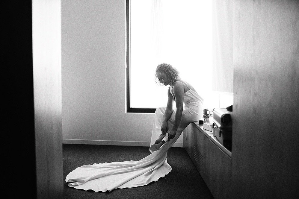 A woman with curly hair in a white dress is sitting on a ledge, putting on her shoes, in a room with a large window and a desk.