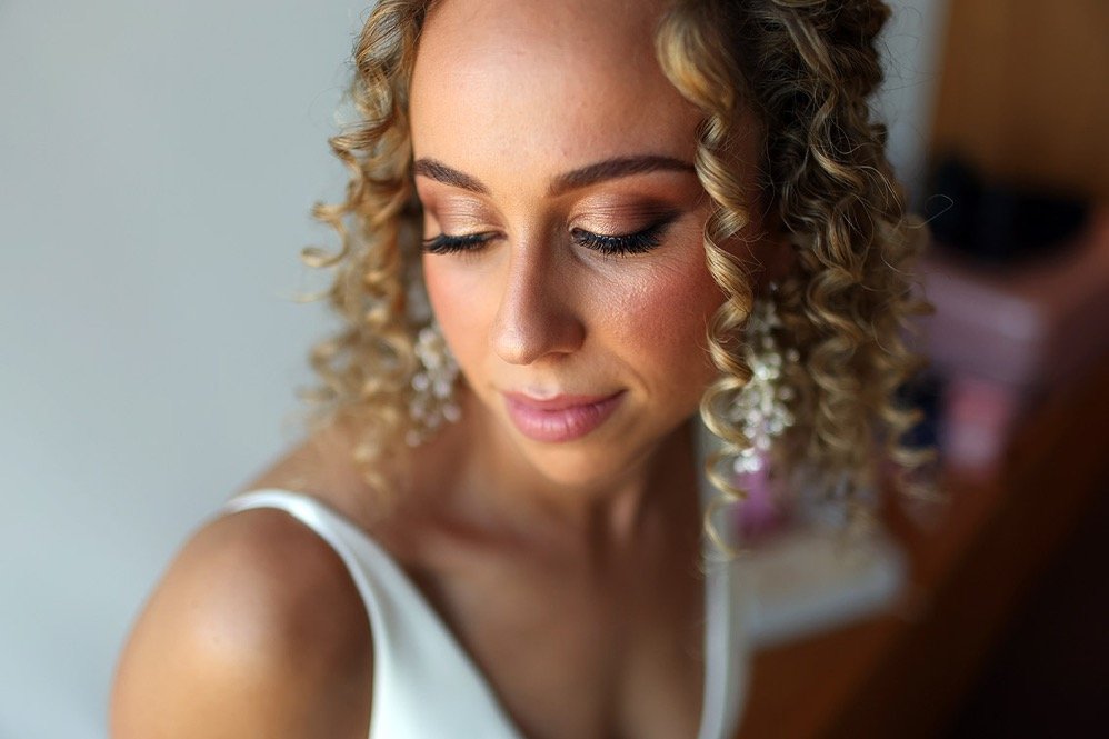Close-up of a woman with curly blonde hair, makeup, and earrings, wearing a white dress, looking down with a gentle smile.