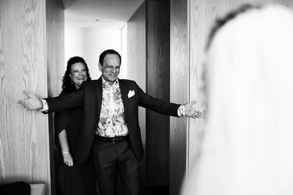 Two people, a woman and a man, smiling and joking in a narrow hallway. The woman is partially hidden behind the man, who has his arms extended outward. The image is in black and white.