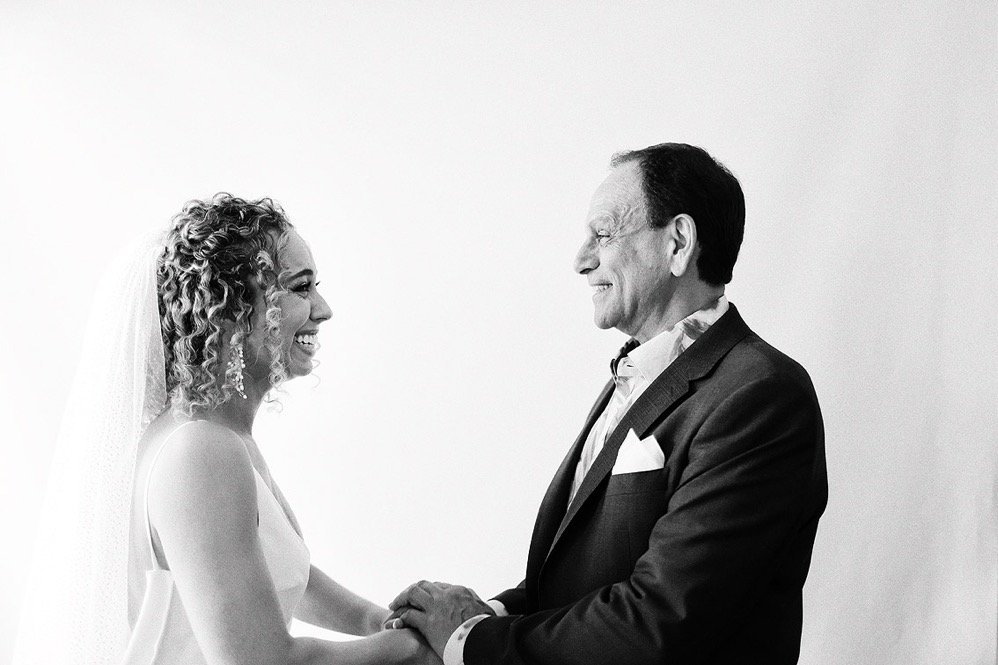 A bride and an older man in a tuxedo holding hands and smiling at each other, likely during a wedding ceremony.