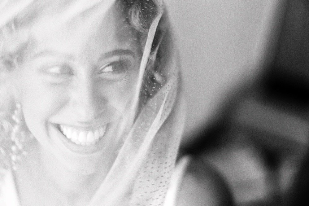 A smiling woman looking through a window or glass with water droplets, possibly on a rainy day.