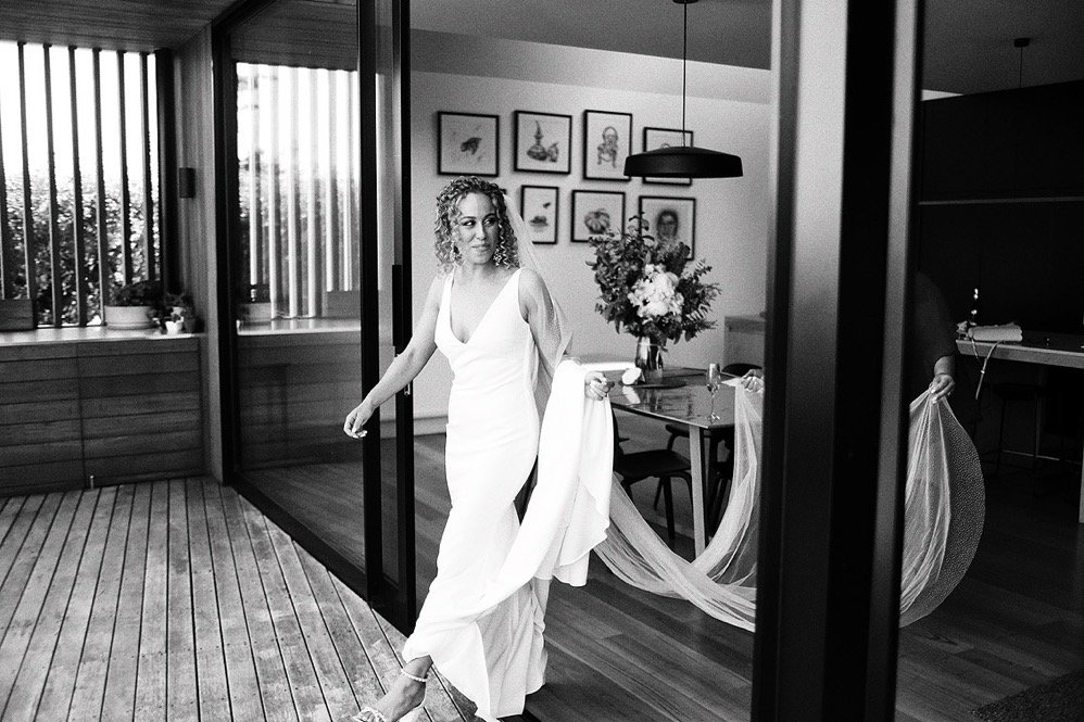 A woman in a white dress walking through a modern dining room. The room has a large flower arrangement on the table, framed art on the wall, and large windows with vertical blinds.