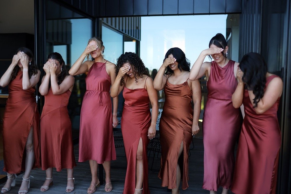 Seven women in matching satin dresses covering their eyes with their hands, standing on a balcony with modern glass buildings in the background.