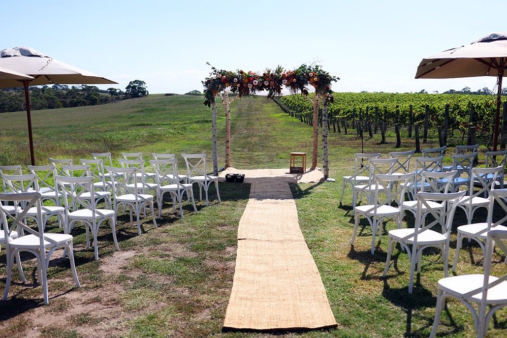 Outdoor wedding ceremony setup with white chairs on either side of a tan aisle runner, decorated arch with flowers, and vineyard in the background under a clear blue sky.