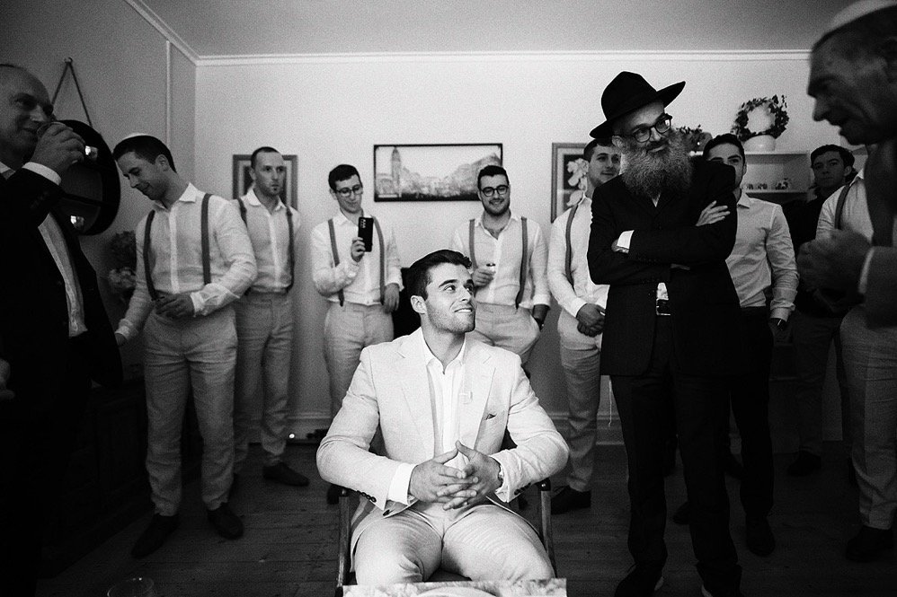 A group of men at a formal event, with one man sitting in a chair wearing a light-colored suit and others standing around him, some taking photos or videos, in a room decorated with framed pictures and a wreath.