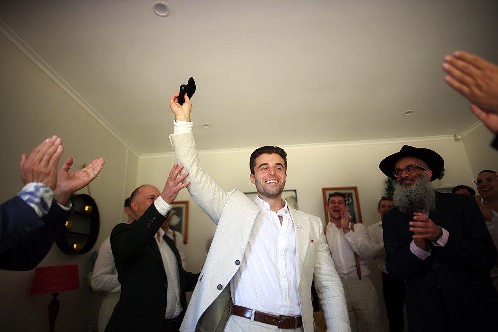 Man in a white suit holding a black shoe, surrounded by people clapping and smiling indoors.