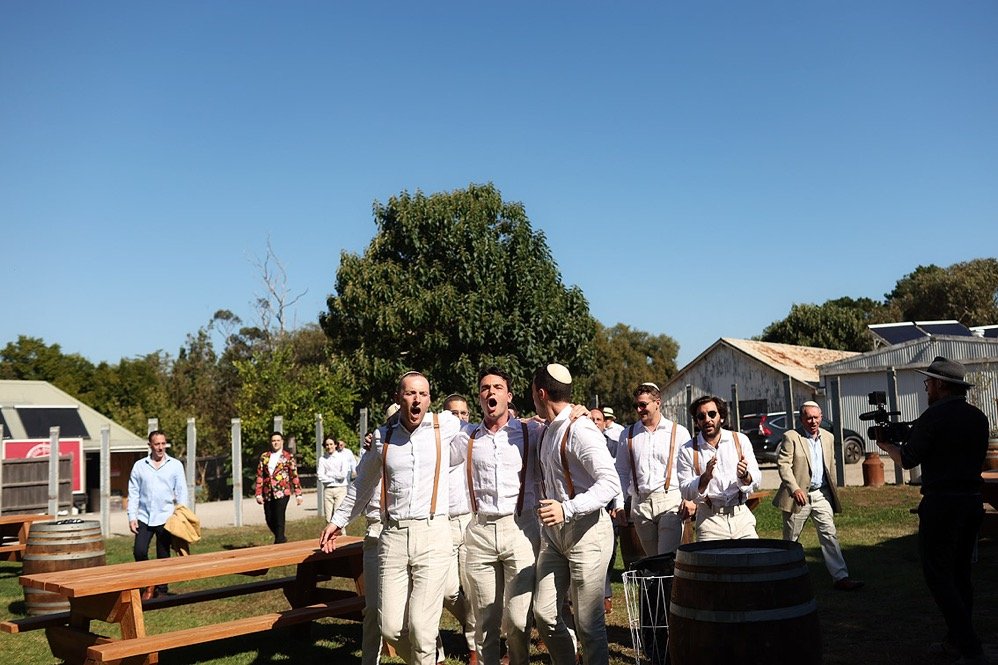 Group of men dressed in vintage style, standing outdoors in a rural setting, with one man speaking into a microphone and holding a tambourine, possibly at a festival or concert.
