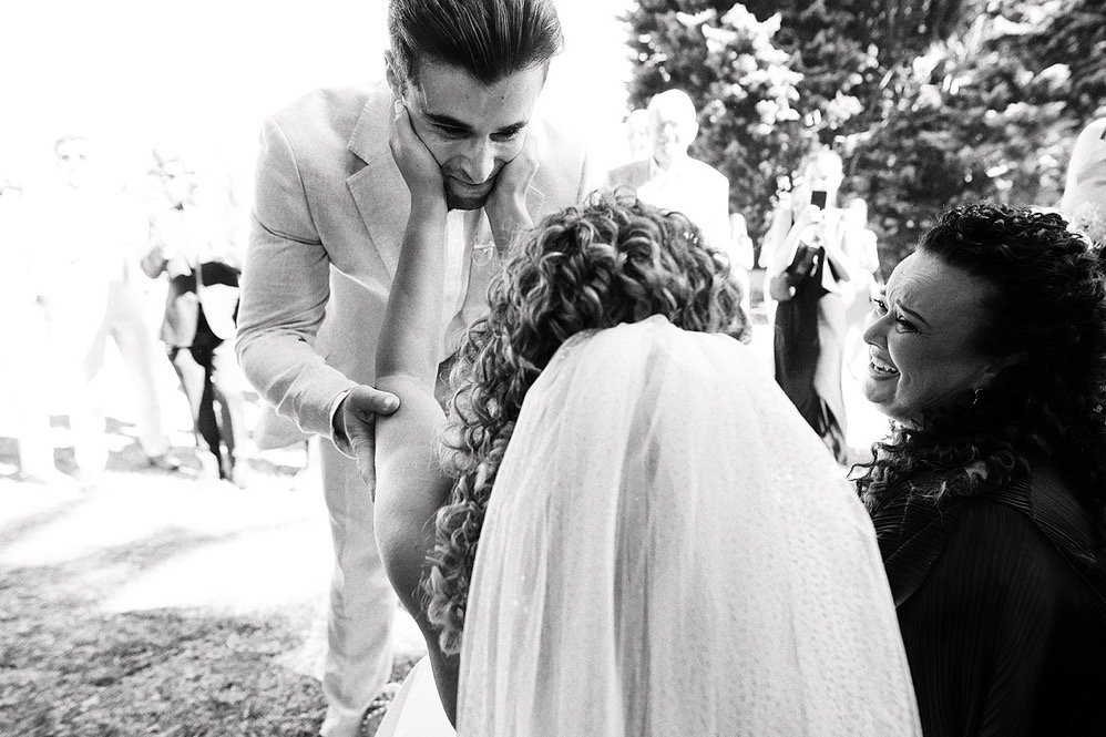 A woman in a wedding dress is hugging and crying as she is comforted by another woman, while a man, possibly the groom, is leaning over and smiling at her. Several people are in the background outdoors.