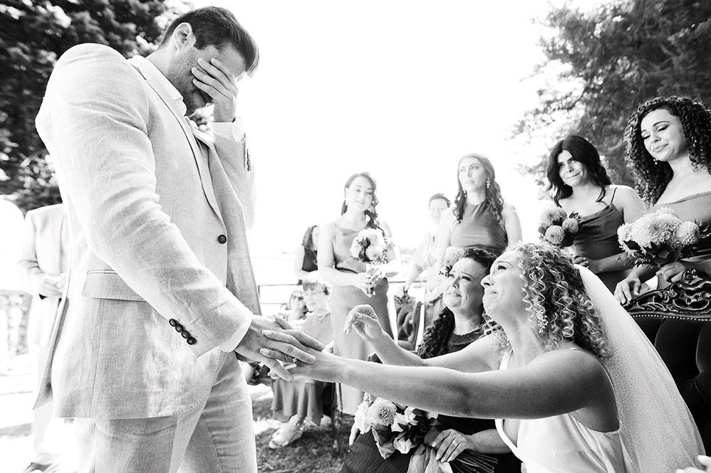 A wedding scene with a bride reaching out her hand to a groom, who is looking down with his hand on his face. The bride is smiling and holding flowers, surrounded by bridesmaids and guests who appear emotional.