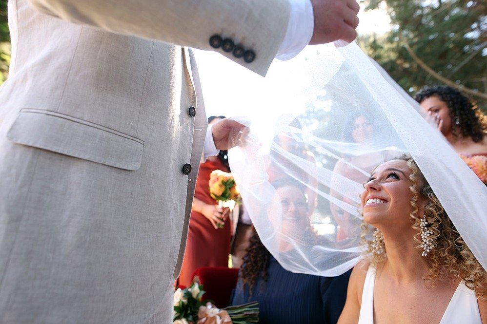 Bride smiling at groom during wedding ceremony, with wedding veil partially covering her face and guests in the background.