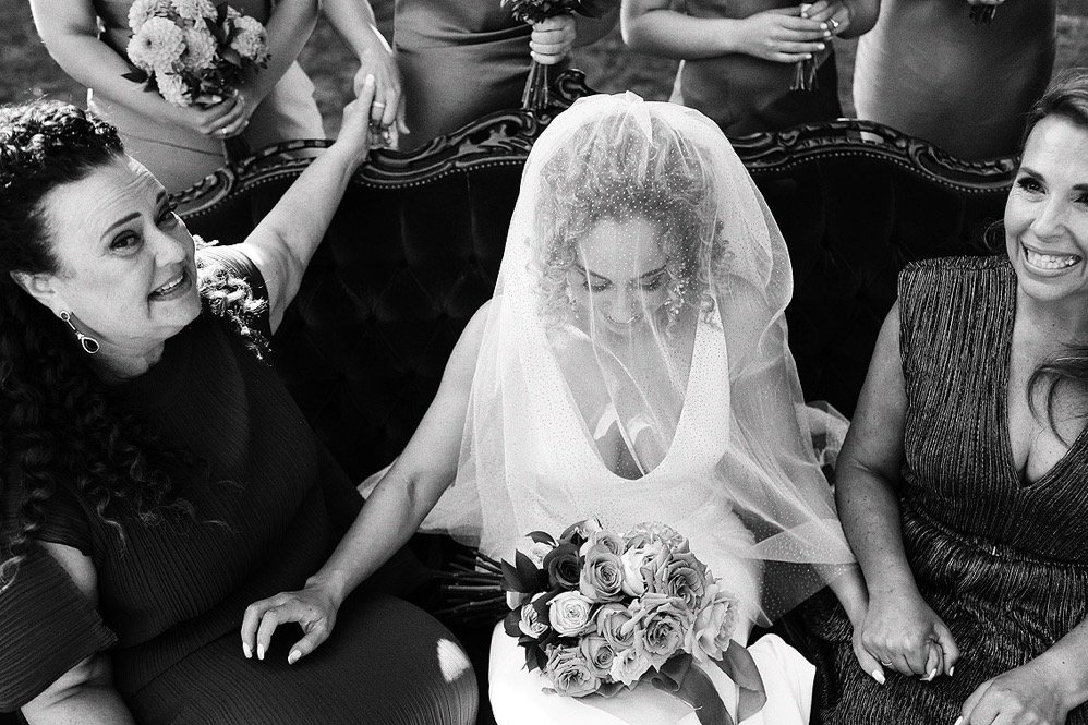 A bride seated on a sofa with her wedding bouquet, surrounded by joyful women, some holding flowers, during a wedding celebration.