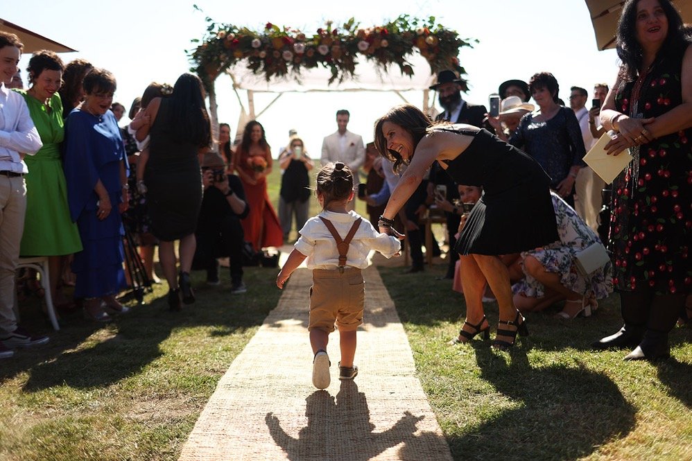 A young child walking down the aisle at an outdoor wedding ceremony, shaking hands with a woman in a black dress, with guests standing on either side and floral decorations overhead.