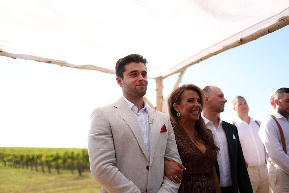 A group of people at an outdoor event, with a man in a white jacket and woman in a brown dress at the center, under a white canopy, with a vineyard in the background.