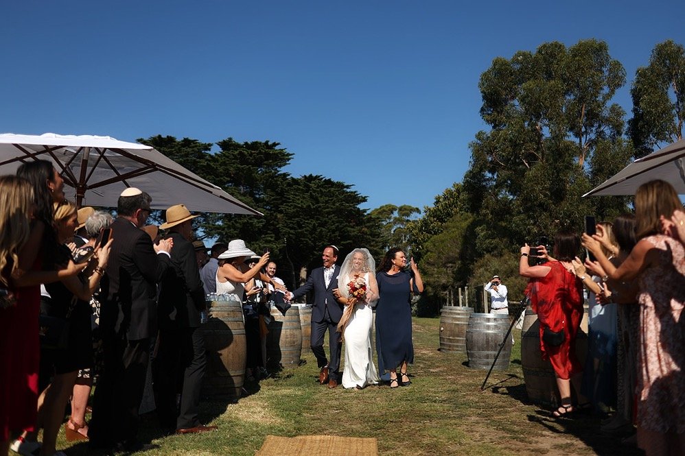 A bride in a white wedding dress holding a bouquet, walking down a grassy aisle with a man in a suit, accompanied by a woman in a navy dress, surrounded by seated and standing guests taking photos and videos, under white umbrellas on a sunny day with
