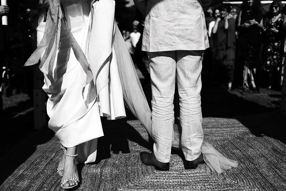 People standing at a wedding ceremony, focusing on the bride's gown and the groom's pants from behind.