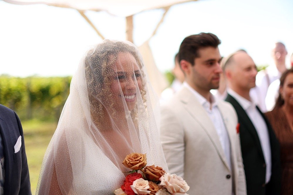 A bride in a wedding veil holding a bouquet of roses smiling during her outdoor wedding ceremony, with groomsmen and guests in the background under a wooden arch.