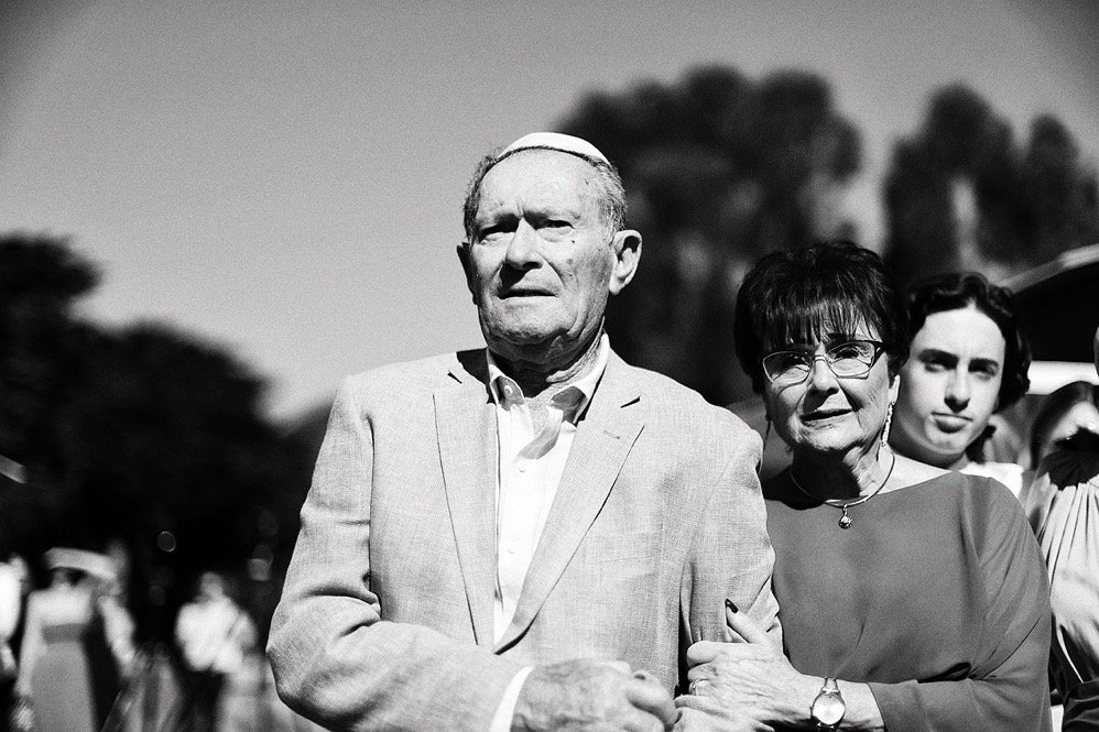An elderly man and woman standing outdoors, holding hands and looking serious, with a young woman in the background.