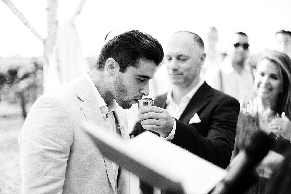Man in light-colored suit drinking champagne at an outdoor event, with other well-dressed people in the background.